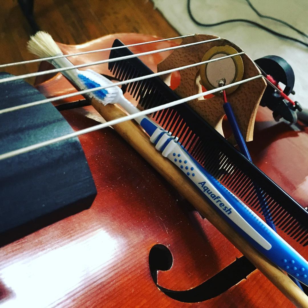 Photo of a cello with a paint brush, toothbrush, comb and contact mic near the bridge